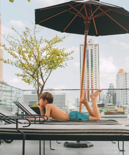 Boy is resting on the roof top under sun umbrella in premium hotel in Bangkok city. Summer holidays